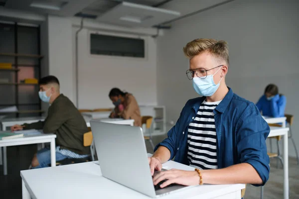 Jovens estudantes com máscaras faciais em mesas na faculdade ou universidade, conceito coronavírus. — Fotografia de Stock