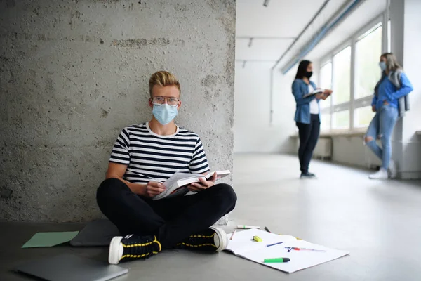 Молодой студент в маске для лица сидит на полу в колледже или университете, коронавирусная концепция. — стоковое фото