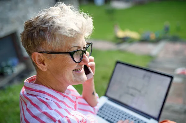 Старша жінка-архітектор зі смартфоном, що працює на відкритому повітрі в саду, концепція домашнього офісу . — стокове фото