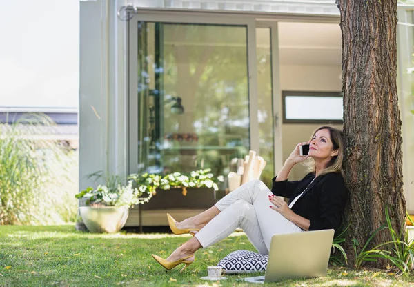 Wanita dewasa yang bekerja di kantor rumah di taman, menggunakan laptop dan smartphone. — Stok Foto