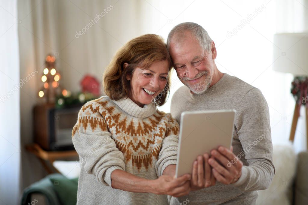 Front view of senior couple with tablet indoors at home at Christmas, taking selfie.