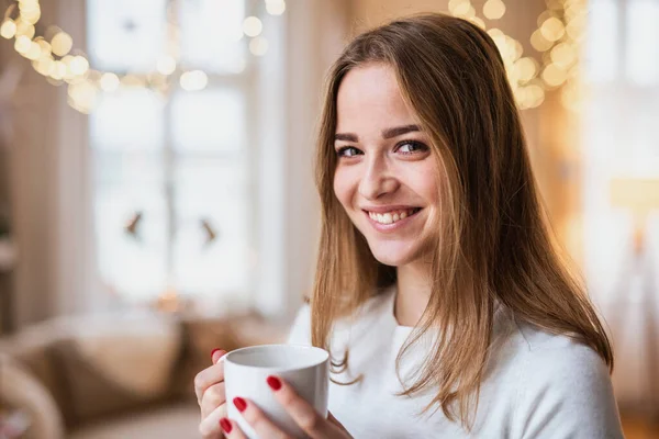 Feliz joven mujer en casa en Navidad, sosteniendo una taza de café. —  Fotos de Stock