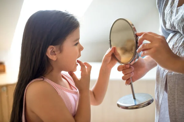 Niña con madre en el baño en el interior, mirando en el espejo en el diente suelto bebé. —  Fotos de Stock