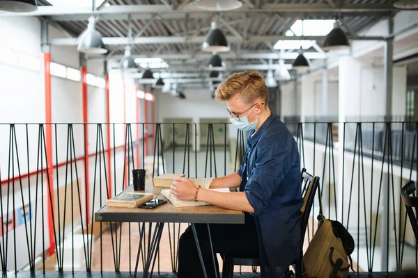 Jonge student met gezichtsmasker aan balie aan universiteit of hogeschool, coronavirus concept. — Stockfoto