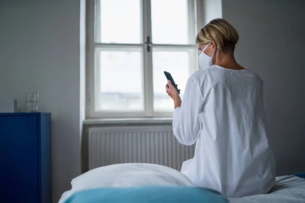 Bakifrån av kvinna patient med smartphone sitter på sängen på sjukhus, psykisk hälsa och coronavirus koncept. — Stockfoto