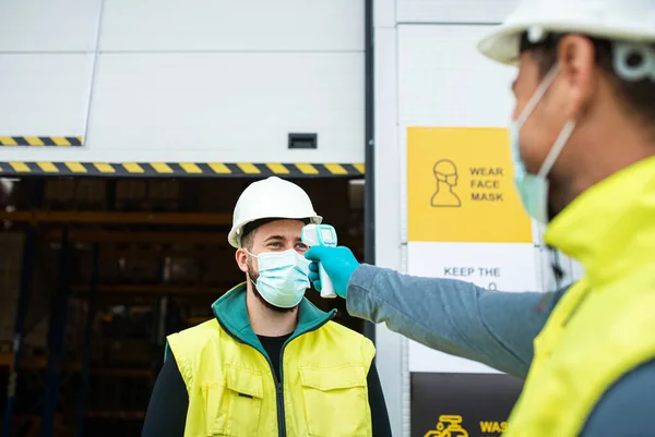 Trabajadores con mascarilla facial frente a almacén, coronavirus y concepto de medición de temperatura. — Foto de Stock