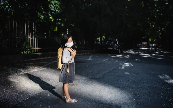 Porträt eines kleinen japanischen Mädchens mit Rucksack, das draußen in der Stadt steht, Coronavirus-Konzept. — Stockfoto