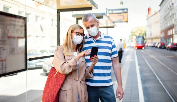 Casal sênior com smartphone em paragem de autocarro ao ar livre na cidade ou cidade, conceito coronavírus. — Fotografia de Stock