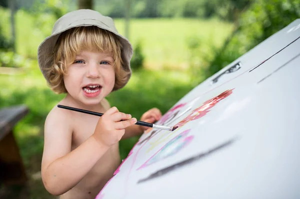 Topless kleine blonde jongen met hoed schilderen buiten in de zomer. — Stockfoto