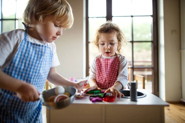 Piccolo ragazzo e ragazza con grembiule giocare al chiuso con cucina giocattolo a casa. — Foto Stock