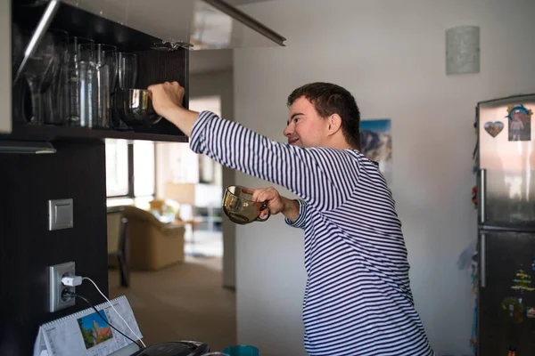 Homme adulte trisomique à l'intérieur dans la cuisine à la maison, aider à la vaisselle. — Photo