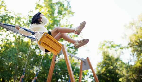 Niña pequeña con mascarilla en swing al aire libre en la ciudad, concepto coronavirus. — Foto de Stock