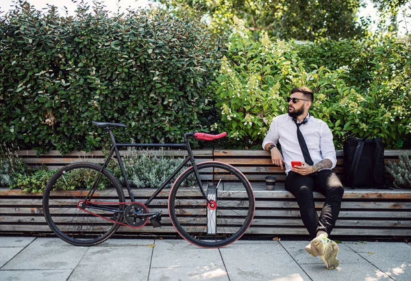 Jovem viajante com bicicleta sentada ao ar livre na cidade, usando smartphone. — Fotografia de Stock