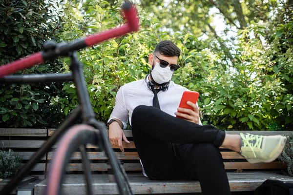 Человек пригородный с велосипедом сидит на открытом воздухе в городе, используя смартфон. Коронавирусная концепция. — стоковое фото