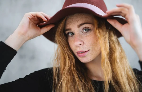 Portret van een jonge vrouw met rood haar en hoed tegen een grijze achtergrond. — Stockfoto