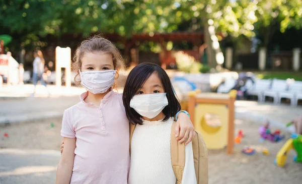 Små skolflickor med ansiktsmask på lekplatsen utomhus i stan, coronavirus koncept. — Stockfoto