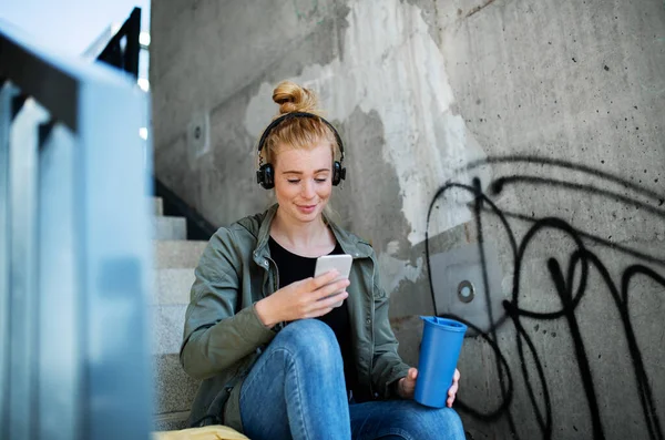 Giovane donna con i capelli rossi all'aperto in città, utilizzando smartphone e cuffie. — Foto Stock