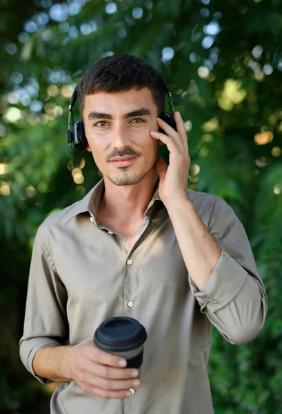 Młody człowiek ze słuchawkami i kawą w kubku wielokrotnego użytku stojącym na świeżym powietrzu w mieście. — Zdjęcie stockowe