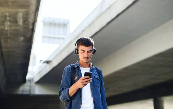 Nízkoúhlý pohled na mladého atraktivního muže se sluchátky stojící venku ve městě. — Stock fotografie