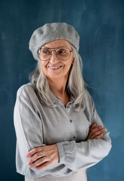 Porträt einer Seniorin mit grauer Baskenmütze, die drinnen vor dunklem Hintergrund steht. — Stockfoto