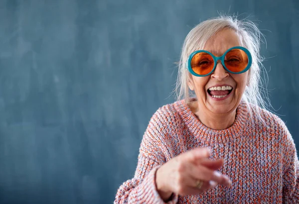 Portrait of senior woman with sunglasses standing indoors against dark background, laughing. — Stock Photo, Image
