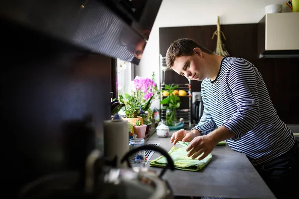 Down syndrom vuxen man står inomhus i köket hemma, hjälper. — Stockfoto