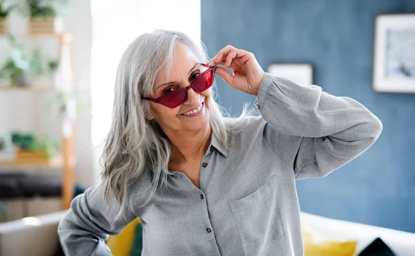 Portrait of senior woman with sunglasses sitting indoors on sofa at home, looking at camera. — Stock Photo, Image