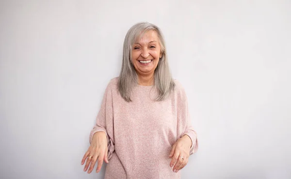 Portret van een oudere vrouw die binnen staat tegen een lichte achtergrond, lachend. — Stockfoto