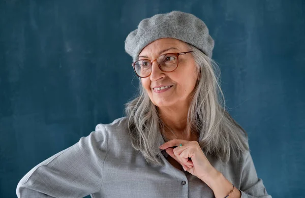 Portret van een oudere vrouw met grijze baret die binnenshuis tegen een donkere achtergrond staat. — Stockfoto