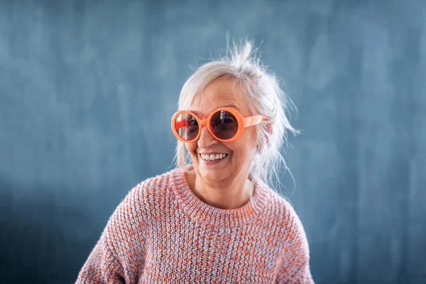Egy idősebb nő portréja napszemüvegben, bent áll sötét háttérrel, nevetve. — Stock Fotó