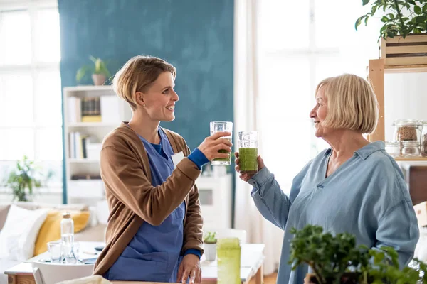 Mulher idosa com cuidador ou profissional de saúde dentro de casa, bebendo smoothie saudável. — Fotografia de Stock