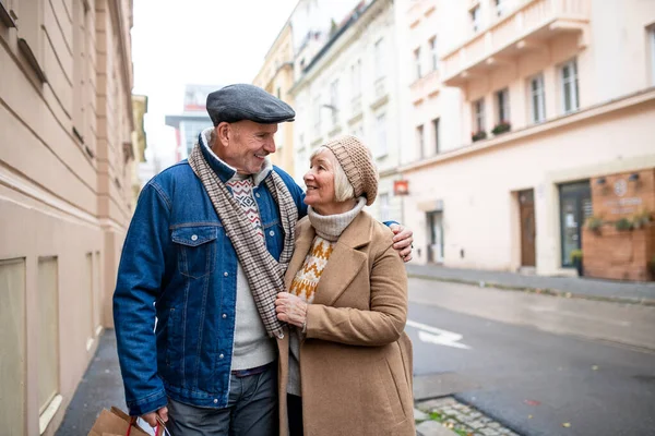 Boldog nyugdíjas pár sétál ki az utcára a városban, bevásárló táskákat cipelve. — Stock Fotó