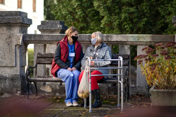 Senior kvinna med promenadram och vårdgivare utomhus sitter i parken, coronavirus koncept. — Stockfoto