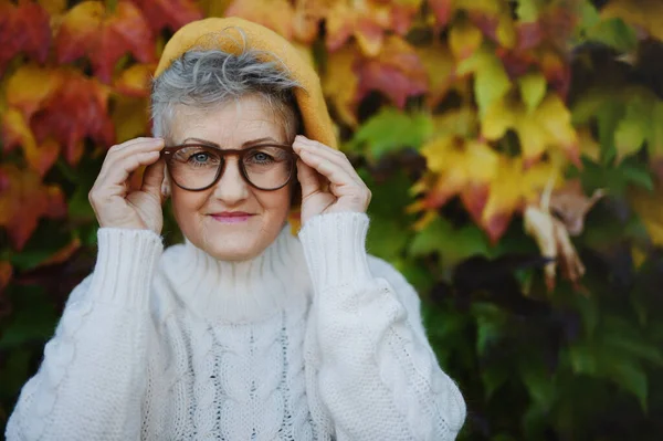 Ηλικιωμένη γυναίκα στέκεται σε εξωτερικούς χώρους κατά πολύχρωμο φυσικό φόντο φθινόπωρο, κοιτάζοντας κάμερα. — Φωτογραφία Αρχείου
