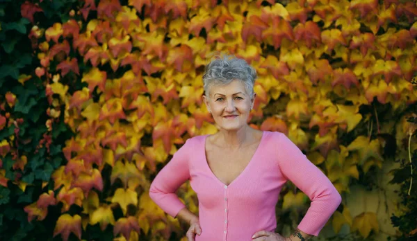 老妇人站在室外，面对着五彩缤纷的秋天背景，看着相机. — 图库照片