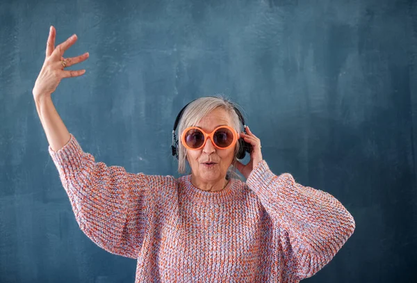Oudere vrouw met een zonnebril en een koptelefoon die binnen tegen een donkere achtergrond staat. — Stockfoto