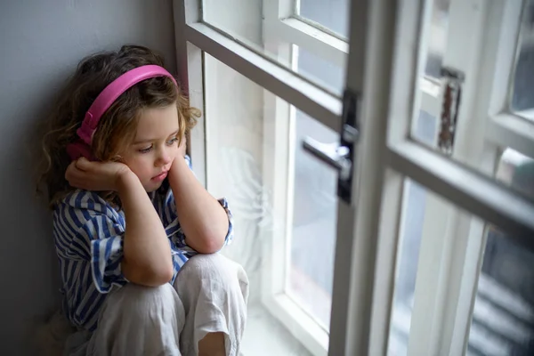 Menina pequena triste com fones de ouvido dentro de casa, sentado no peitoril da janela. — Fotografia de Stock