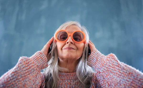 Portret van een oudere vrouw met een zonnebril die binnen tegen een donkere achtergrond staat en naar muziek luistert. — Stockfoto