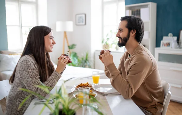 Νεαρό ερωτευμένο ζευγάρι τρώει χάμπουργκερ σε εσωτερικούς χώρους στο σπίτι. — Φωτογραφία Αρχείου