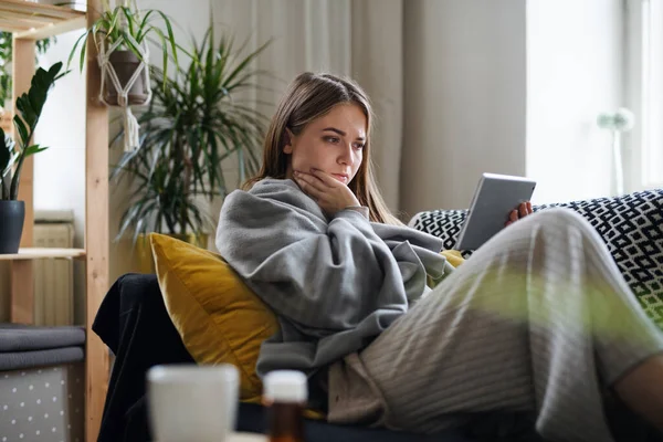 Ill young woman wrapped in blanket on sofa at home, using tablet. — Stock Photo, Image