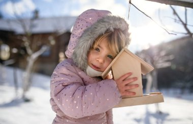 Kışın bahçede küçük bir kız, tahta kuş yemliğinin yanında duruyor..