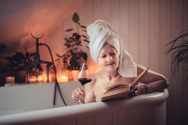 Glückliche Seniorin liest Buch und trinkt zu Hause Wein in Badewanne. — Stockfoto