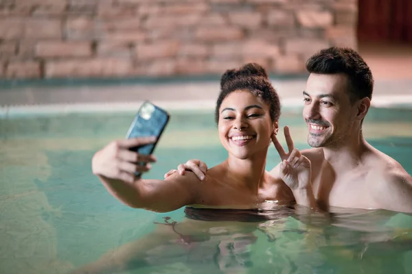 Retrato de jovem casal em piscina termal interior de fonte termal, tirando selfie. — Fotografia de Stock