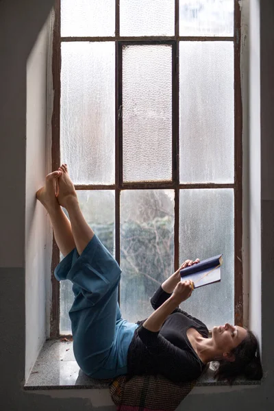 Mulher deitada no peitoril da janela velha e suja, lendo um livro. — Fotografia de Stock