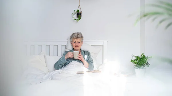 Glad äldre kvinna med kaffe läsa bok i sängen hemma, tittar på kameran. — Stockfoto