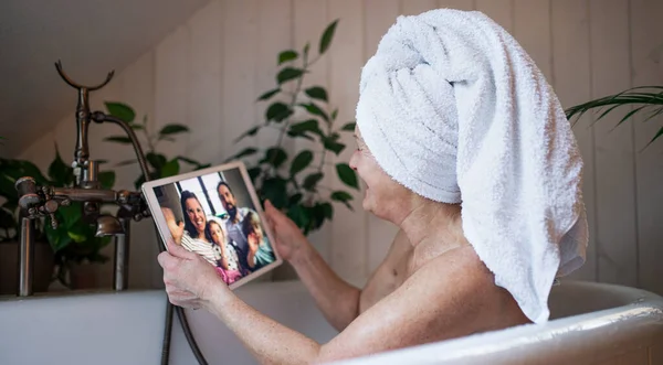 Femme âgée avec tablette dans la baignoire à la maison, appel vidéo avec fille et petits-enfants. — Photo