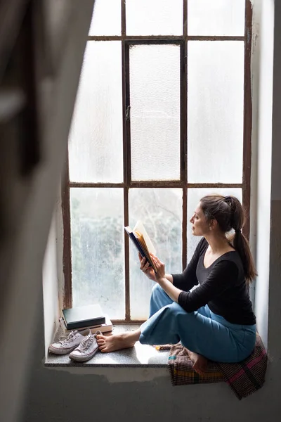 Mutlu kadın eski ve kirli pencere pervazında oturmuş kitap okuyor.. — Stok fotoğraf