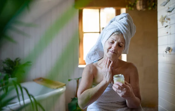 Felice donna anziana avvolta in un asciugamano in bagno a casa, applicando idratante corpo fatto in casa. — Foto Stock
