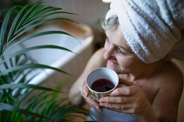 Ευτυχισμένη ηλικιωμένη γυναίκα τυλιγμένη σε πετσέτα στο μπάνιο στο σπίτι, να ξεκουράζεται με τσάι.. — Φωτογραφία Αρχείου