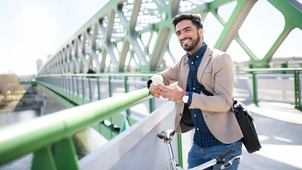 Jonge zakenman forens met fiets naar buiten te werken in de stad, met behulp van de smartphone op de brug. — Stockfoto